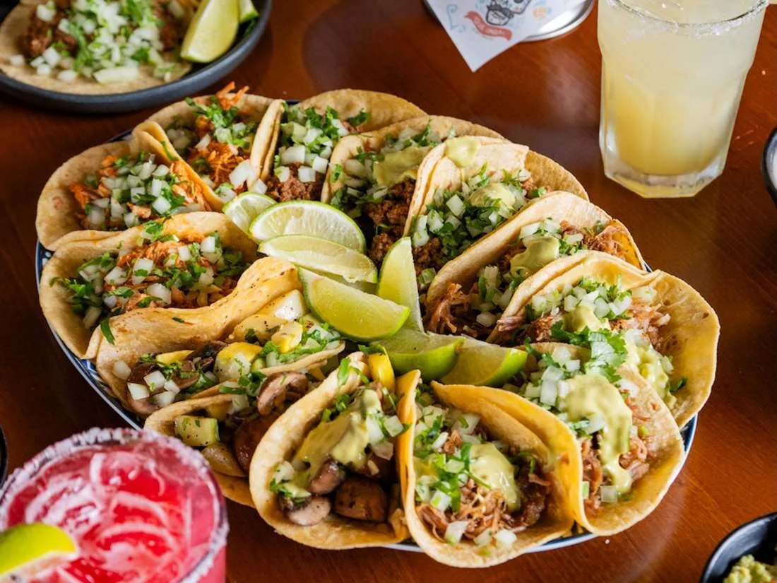 A plate of assorted tacos filled with meat, topped with chopped onions and cilantro, served with lime wedges. Also includes a glass of a yellow beverage with ice and lemon slices, and a pink drink with a lime wedge in a glass with a salt rim.