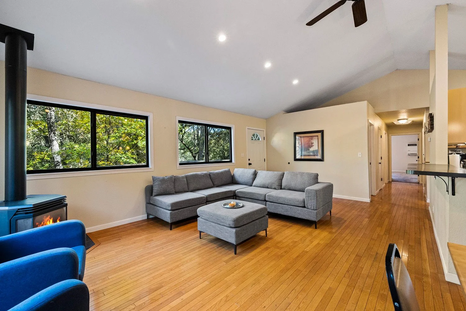 Living room with a gray sectional sofa, wooden flooring, and large windows showing trees outside. There is a fireplace with a fire, a framed picture on the wall, and a hallway leading to other rooms.