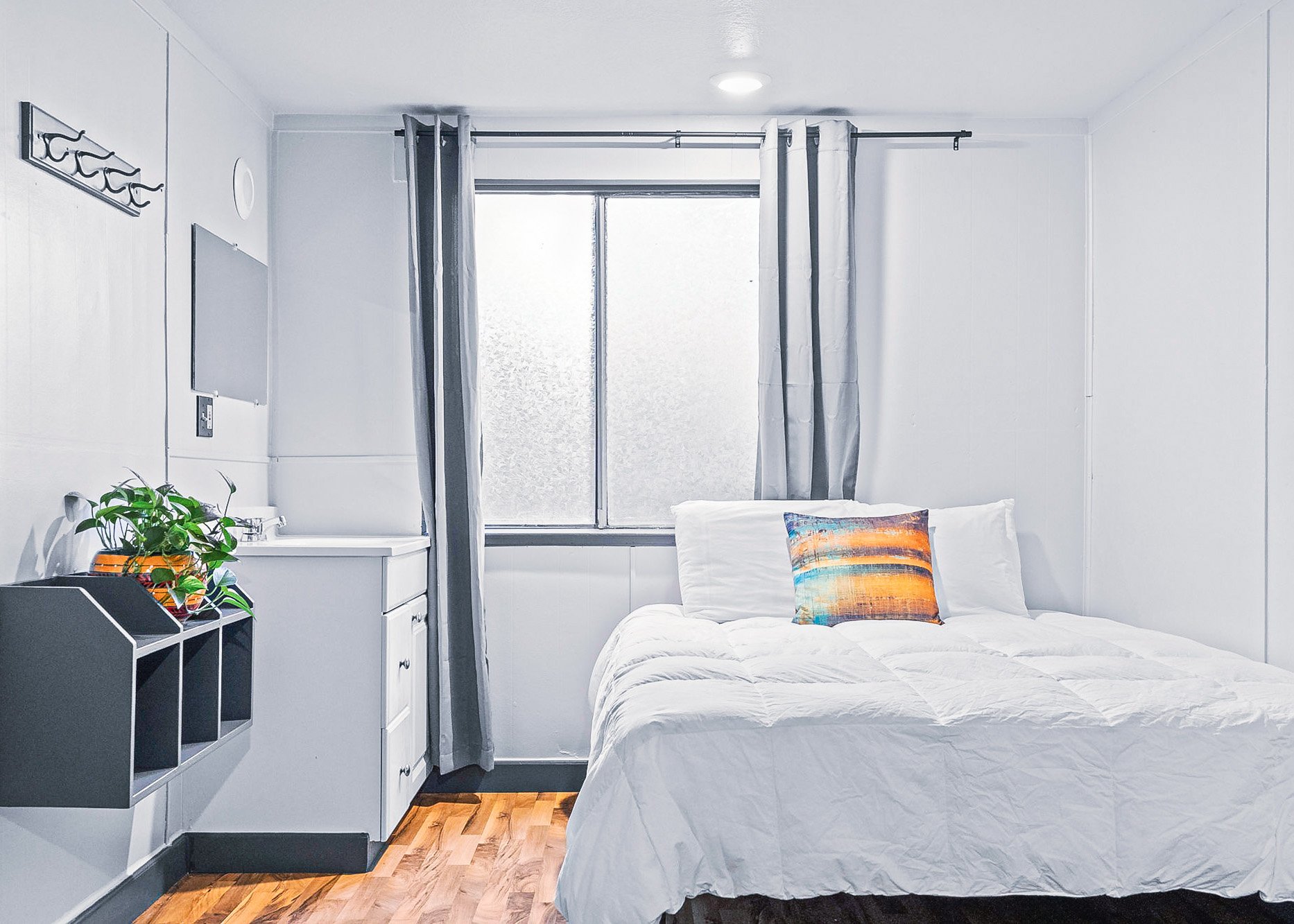 A bright, minimalistic lodge room with white walls and bed, a colorful pillow, a window with gray curtains, a small shelf with plants and books, and a wall-mounted coat rack.