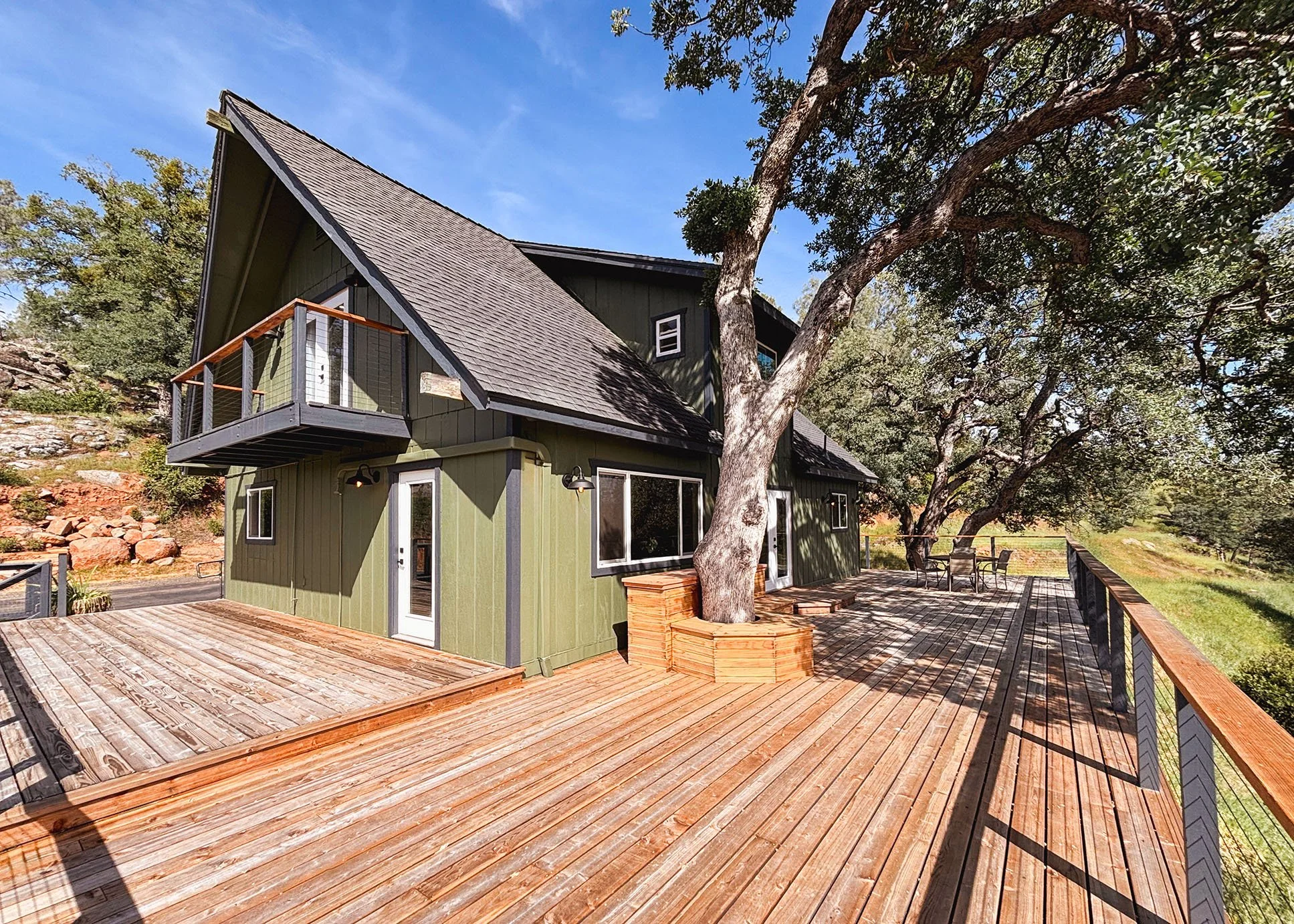 A green house with a shingled A-frame roof and multiple windows, featuring a small balcony with a wooden railing. There is a large tree with multiple trunks growing close to the house, casting shadows on the exterior. The house has a spacious wooden deck with outdoor chairs and a table, surrounded by a natural landscape with trees and rocks under a clear blue sky.