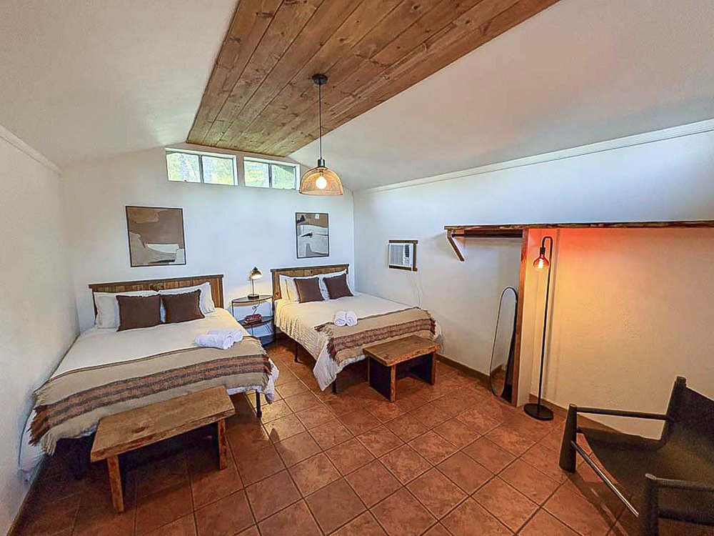 A cozy cabin with two beds, a small wooden bench at the foot of each bed, wall art, a shared nightstand with a lamp, a window AC unit, a floor lamp with an exposed bulb, a mirror, a sitting chair, and a wooden ceiling with a hanging light fixture.