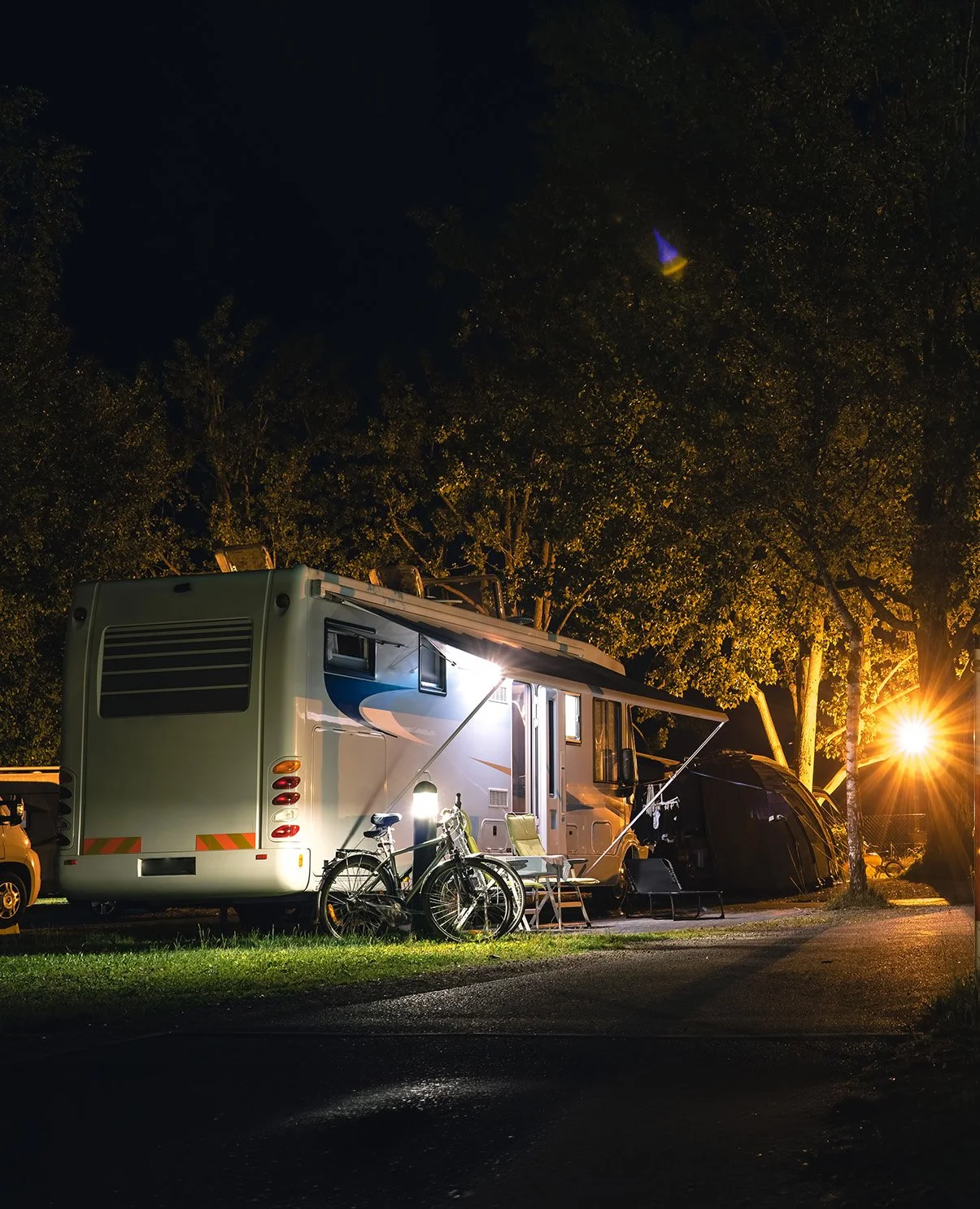 Night scene of a recreational vehicle parked with bicycles and outdoor chairs under trees, illuminated by outdoor lights.