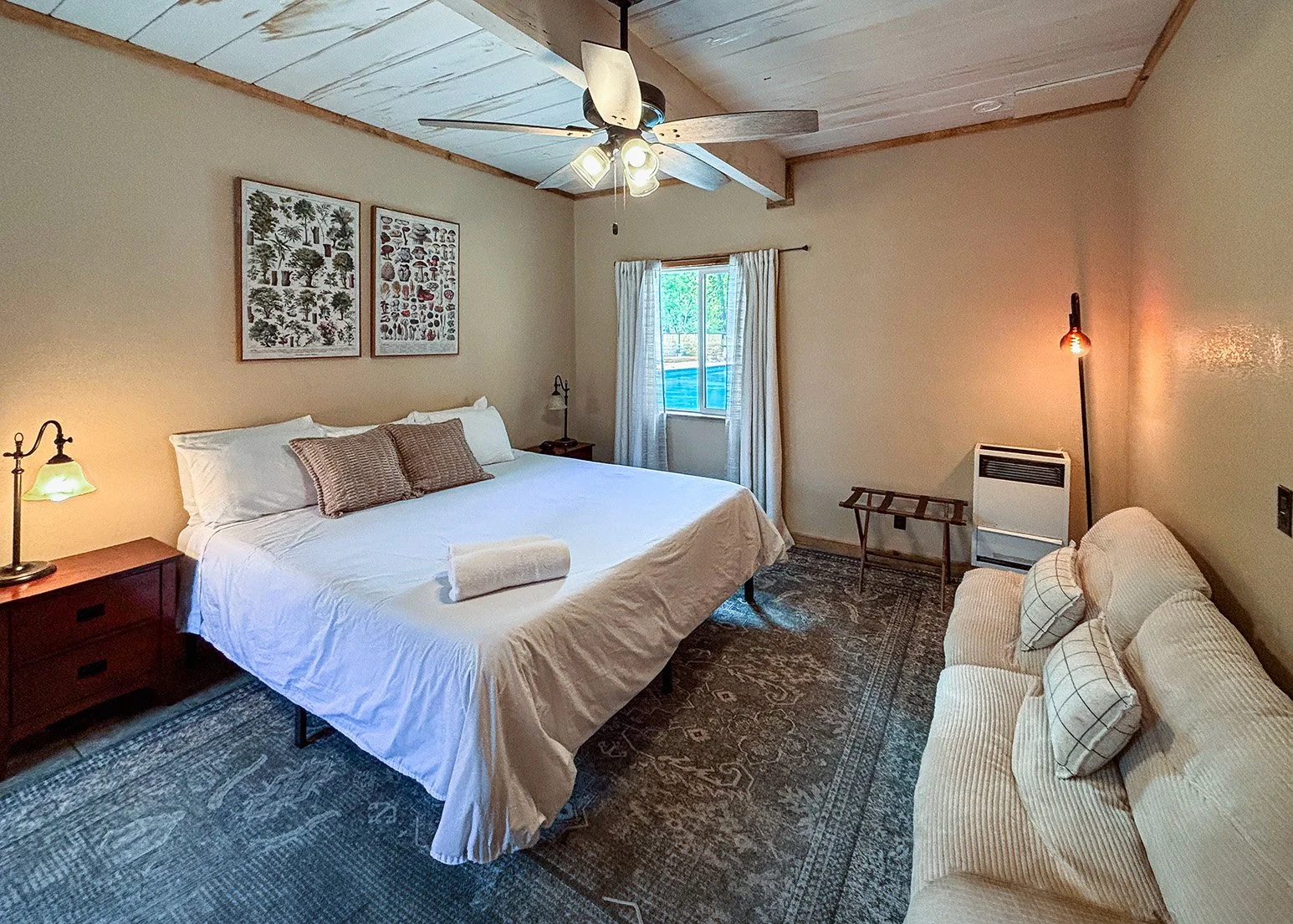 A cozy cabin with a large bed covered in white linens, pillows. A nightstand with a lamp on each side of the bed. The room has beige walls, a window with white curtains, and a ceiling fan. A beige sofa with checkered pillows is on the right.