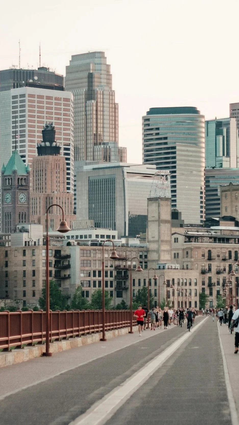 City skyline with tall buildings and a mix of historical and modern architecture, viewed from a pedestrian bridge with people walking and biking.