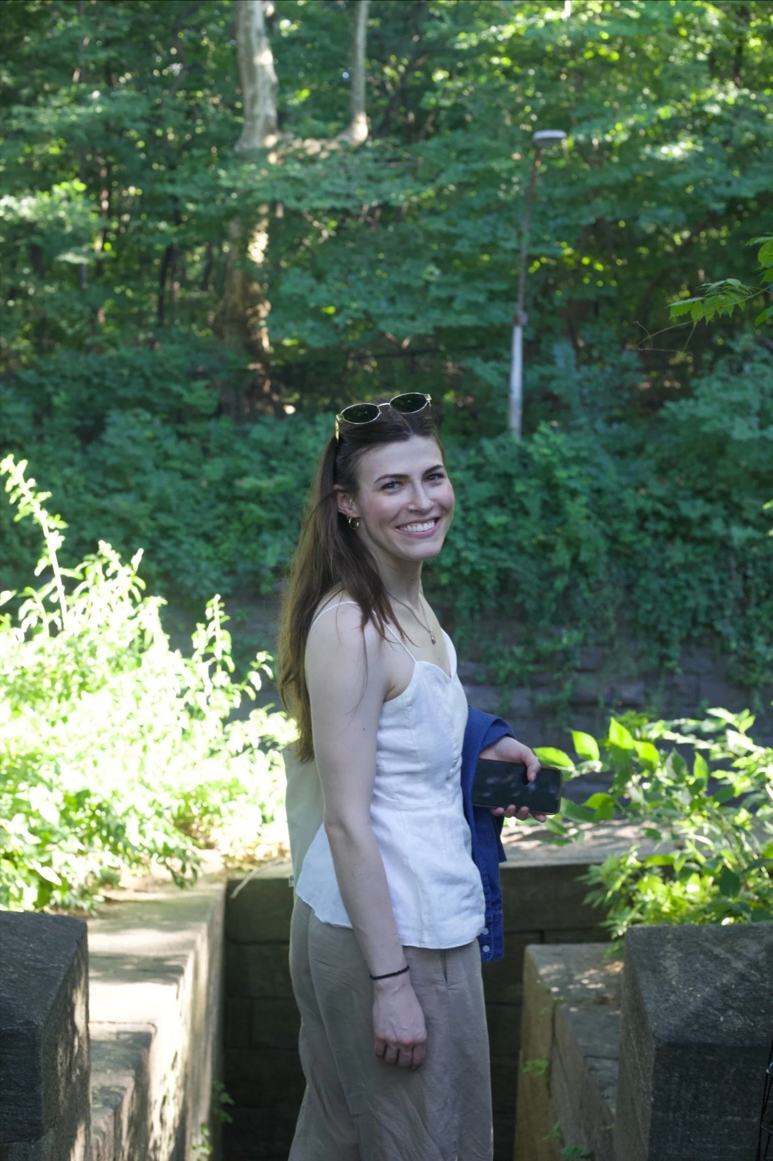 A woman with long brown hair, sunglasses on her head, wearing a light-colored sleeveless top and tan pants, smiling and holding a smartphone, standing on a stone pathway in a lush green park with trees and bushes surrounding her.
