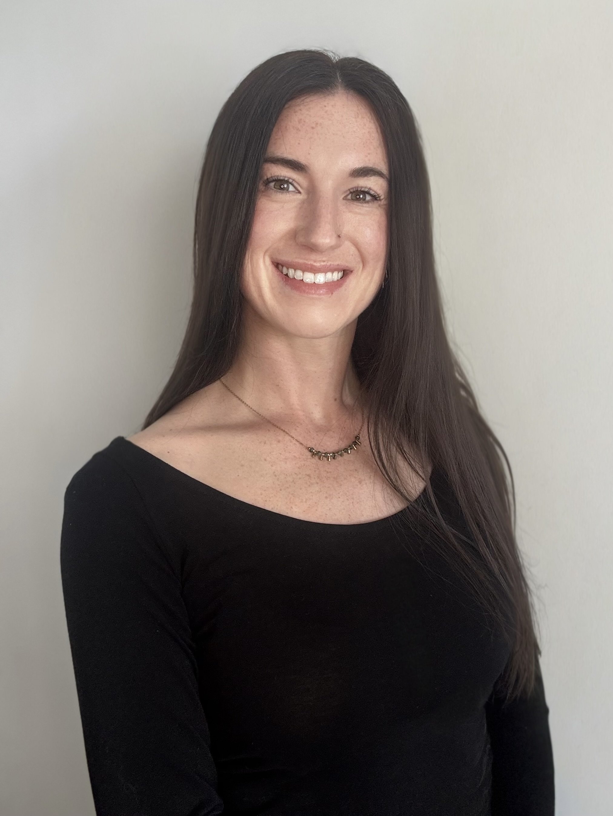 A woman with long brown hair, light skin, and freckles, smiling with visible teeth. She is wearing a black top, and a necklace, standing against a plain light-colored wall.