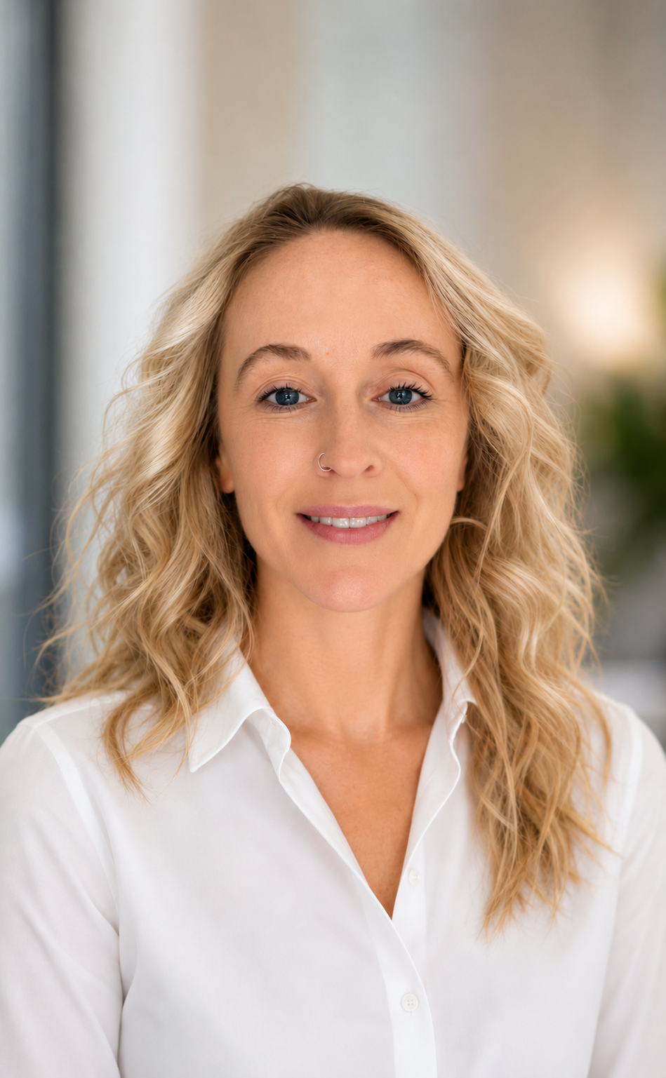 A woman with curly blonde hair, blue eyes, and a nose piercing, wearing a white button-up shirt, smiling at the camera.