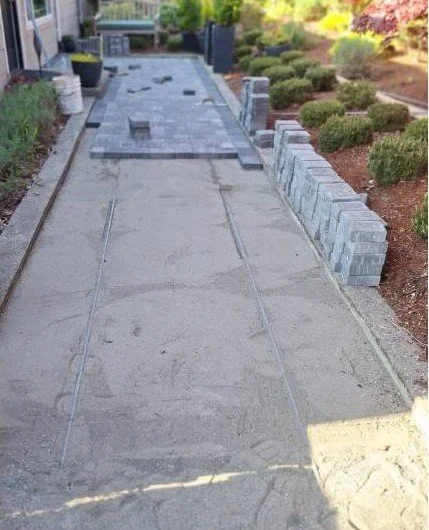 A sidewalk under construction with concrete sections and some stacked bricks on the right, surrounded by shrubbery and residential buildings.