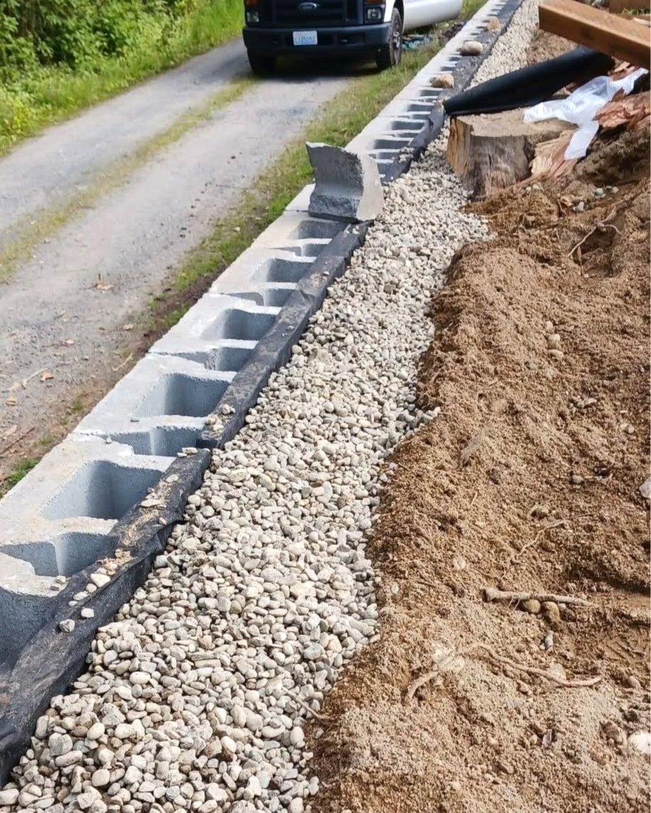 Close-up of a construction site showing a partially completed concrete block wall with gravel and dirt nearby. A black plastic sheet lines part of the ground, and a large piece of concrete block is resting on the wall. In the background, a pickup tru