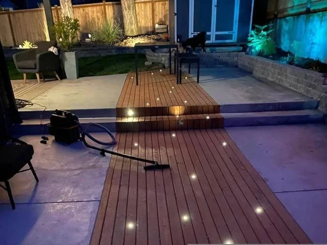 Backyard patio with a wooden deck illuminated by small embedded lights, a vacuum cleaner in the foreground, and outdoor furniture against a fenced yard with plants and lighting.