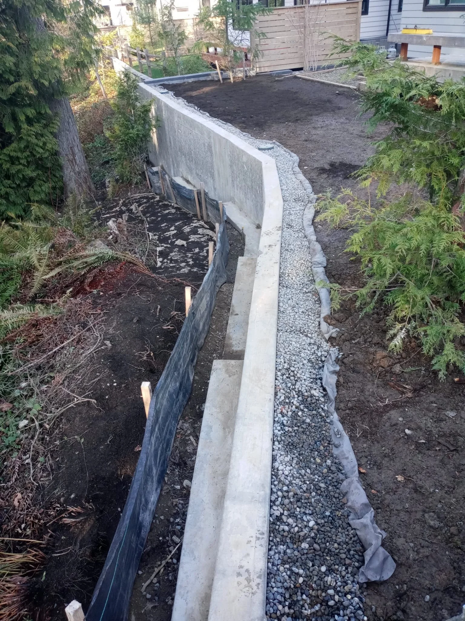 A landscaped backyard with a newly poured concrete garden bed, black plastic edging with soil and plants, and a gravel pathway along the side.