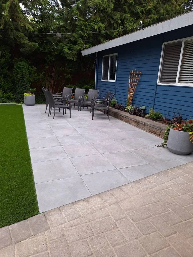 Patio with outdoor dining chairs, concrete tiles, potted plants, and a blue house with windows, surrounded by greenery.