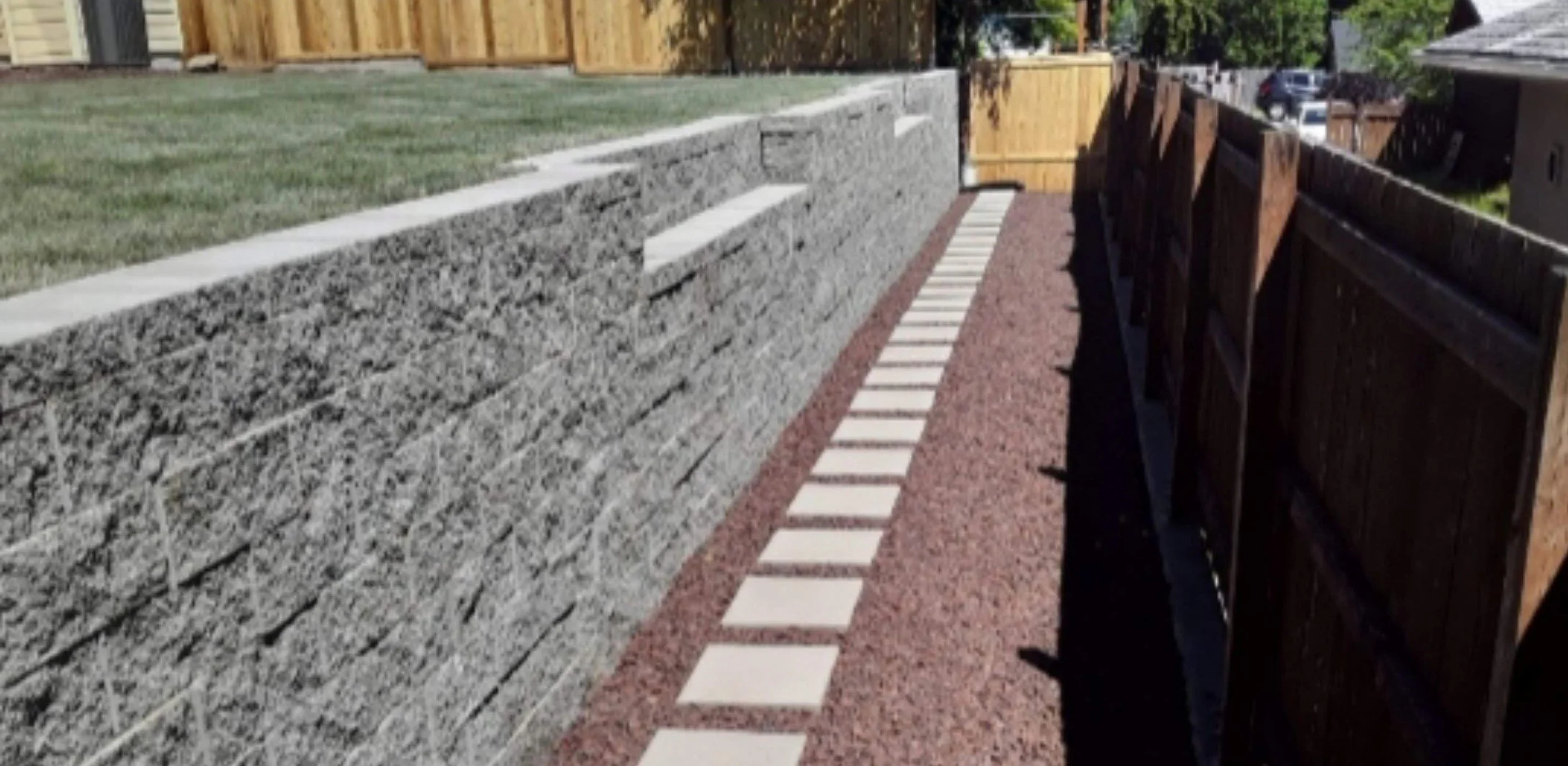 A backyard with a gravel pathway bordered by white stepping stones, a stone retaining wall with concrete caps, and a wooden fence on the right side.