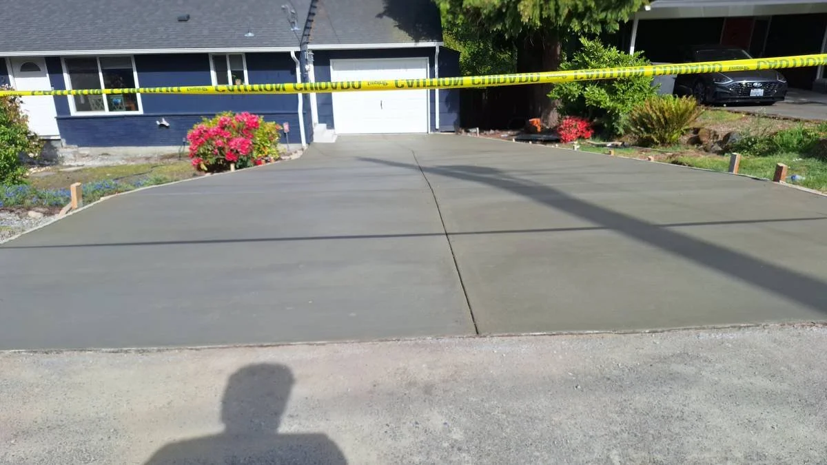 Freshly poured concrete driveway with caution tape in front of a house with blue exterior and flower beds, indicating recent construction or repair.