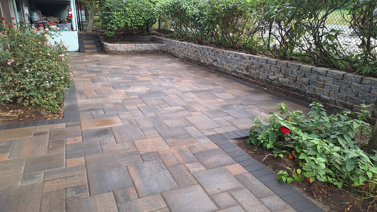 A landscaped backyard patio with brick and stone pavers, bordered by a low stone wall and garden beds with bushes and flowers.