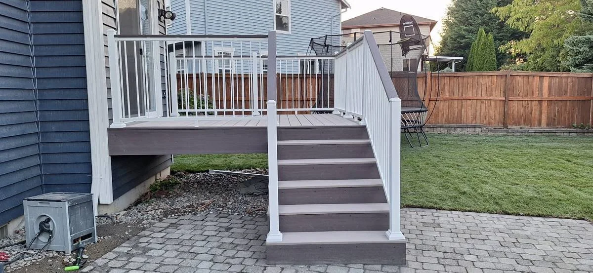 Backyard with a raised wooden deck, white railings and stairs leading down to a stone patio. There is a grassy area beyond the patio, enclosed by a wooden fence, with outdoor chairs and trees in the background.