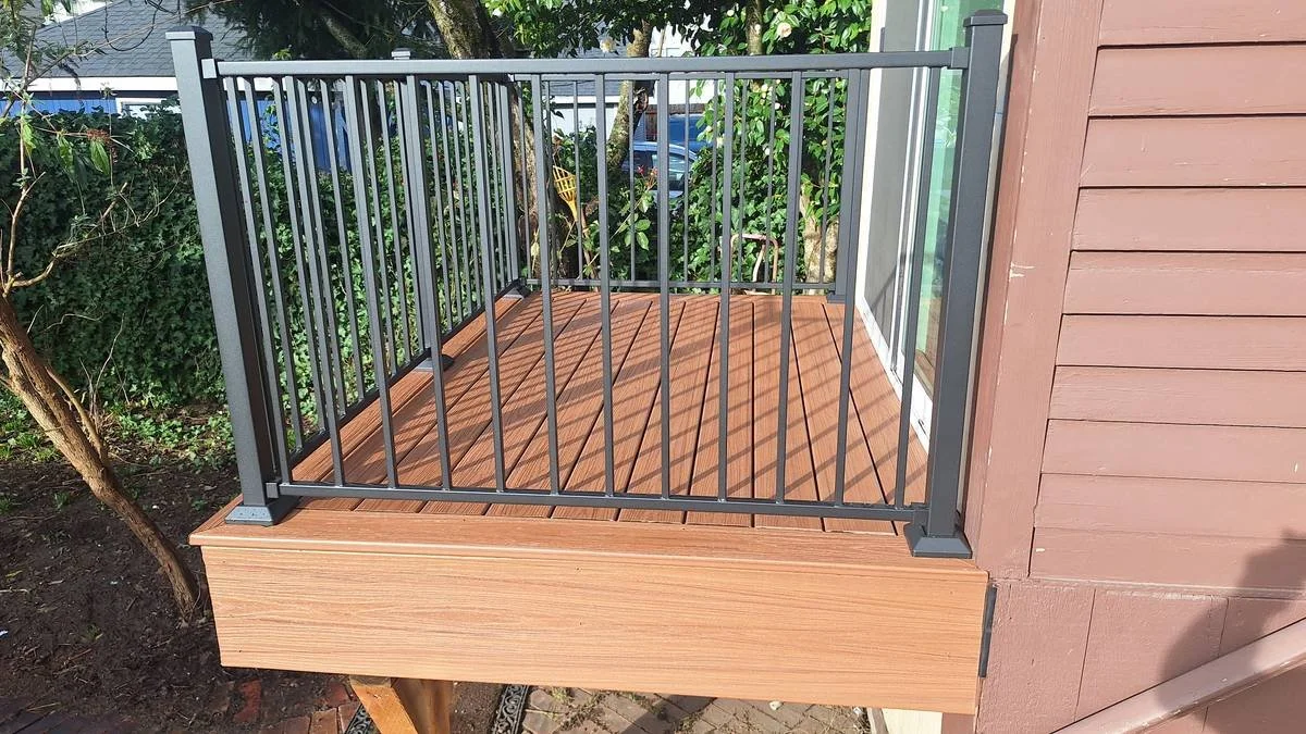 A small outdoor wooden deck with a metal railing, attached to a house with brown siding, surrounded by green bushes and trees.