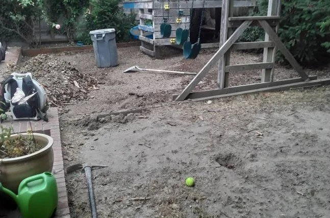 A backyard with a dirt area, a green watering can, a gardening shovel, a large plant pot, a trash can, and a raised garden bed with gardening tools hanging on it.