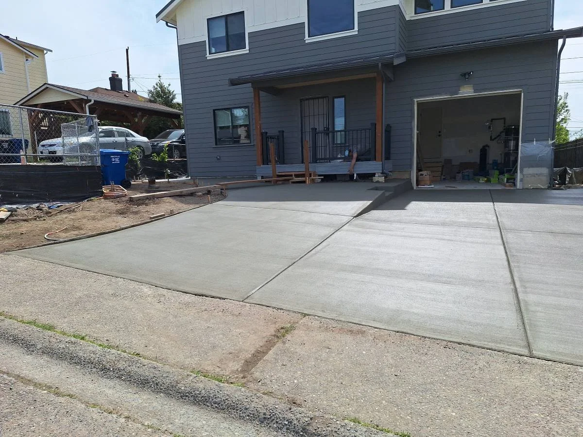 Concrete driveway in front of a residential house under construction, with some tools and materials nearby, and a garage door open revealing a partially finished interior.