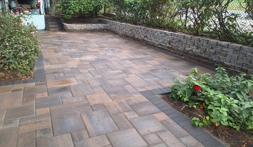 A paved backyard patio with a raised garden bed on the right, a brick wall on the back, and a stone steps leading up at the back left corner.