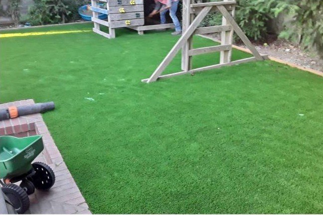 A backyard with green artificial turf, a small wooden play structure, a child's green wagon, and a brick patio area.