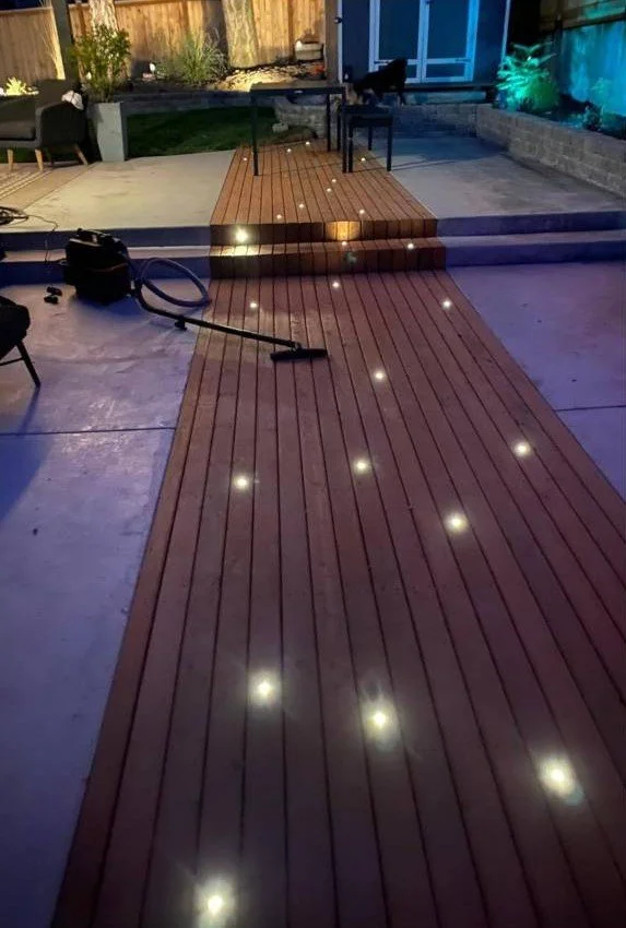 Backyard deck with lights illuminating the wooden planks, vacuum cleaner on the concrete, and outdoor furniture including a table and chairs.