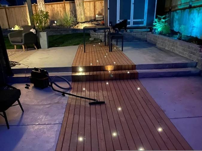 Backyard patio with wooden deck, embedded lights, outdoor furniture, a vacuum cleaner on the deck, and a dog near the steps at night.