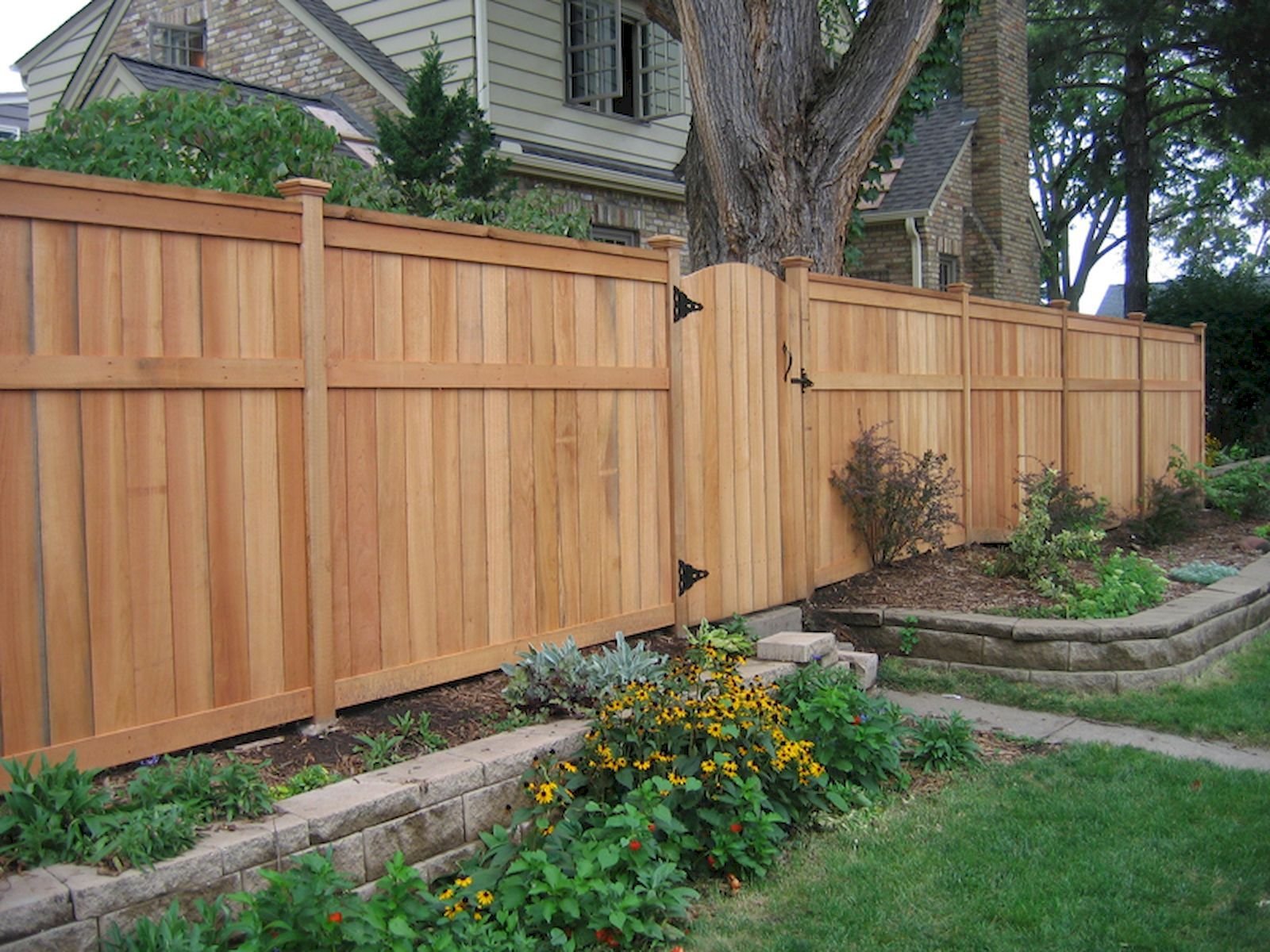 A newly installed wooden fence in front of a garden with plants and flowers, alongside a large tree and a brick house in the background.