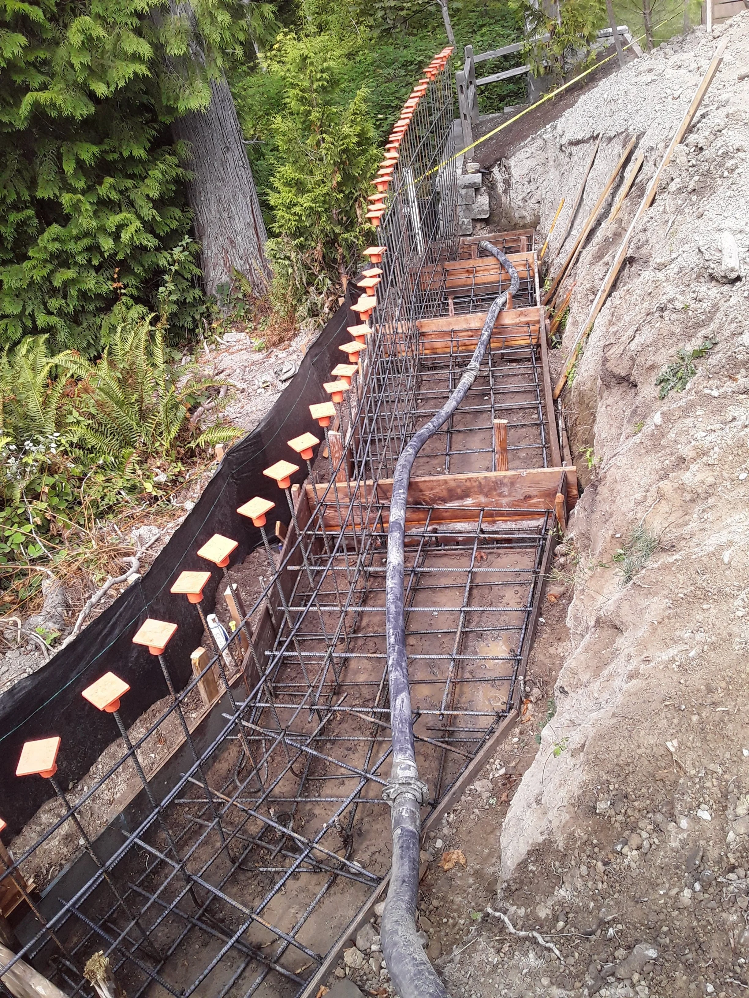 Construction site with rebar framework, wooden planks, and pipes in a trench, surrounded by trees and rocks.