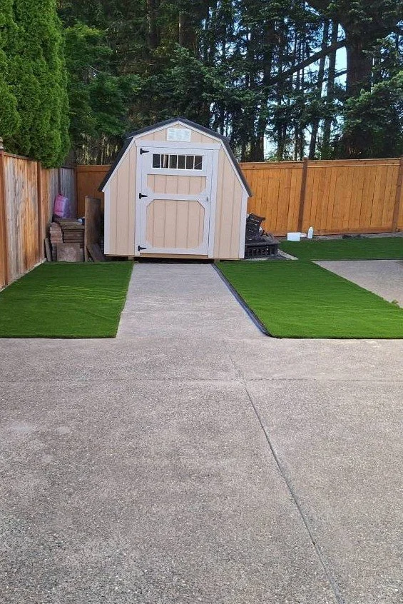 Backyard with a beige garden shed, wooden fence, concrete ground, and patches of artificial grass by Kairos Hardscape & Design