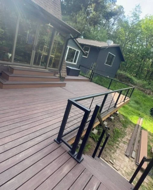 A wooden deck attached to a house, with black metal railings, overlooking a grassy yard with trees.