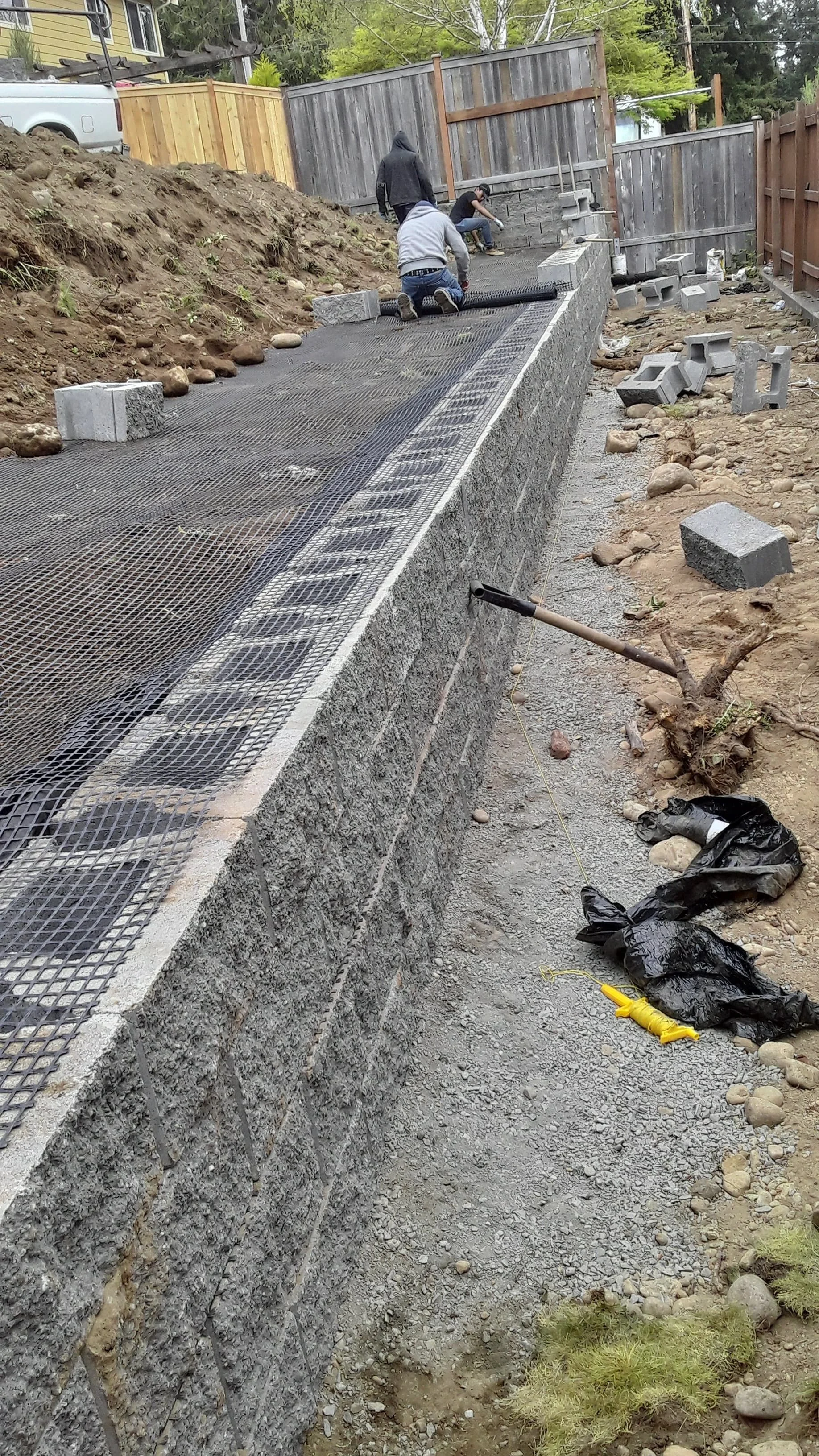 Workers are constructing a stone retaining wall with concrete cinder blocks and wire mesh fencing. They are laying rebar and working on leveling the area, surrounded by construction materials, dirt, and rocks.