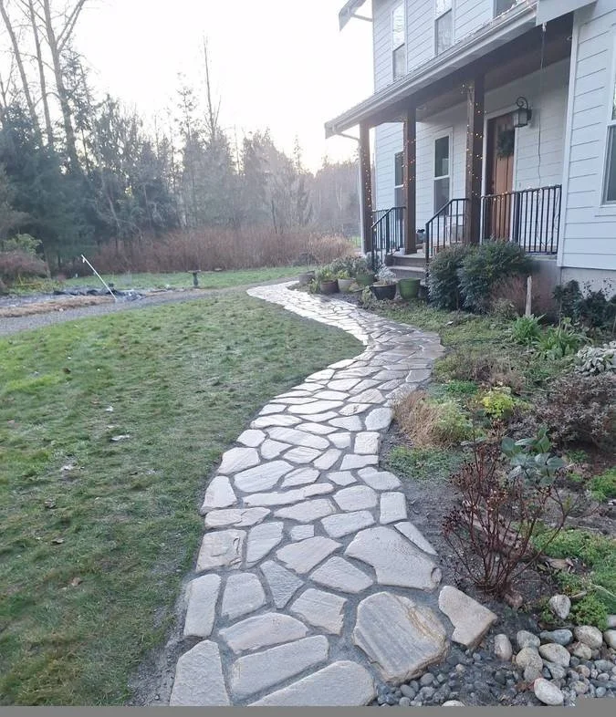 Curved stone pathway leading to the front porch of a house with black railing, surrounded by a lawn and garden with potted plants and bushes.