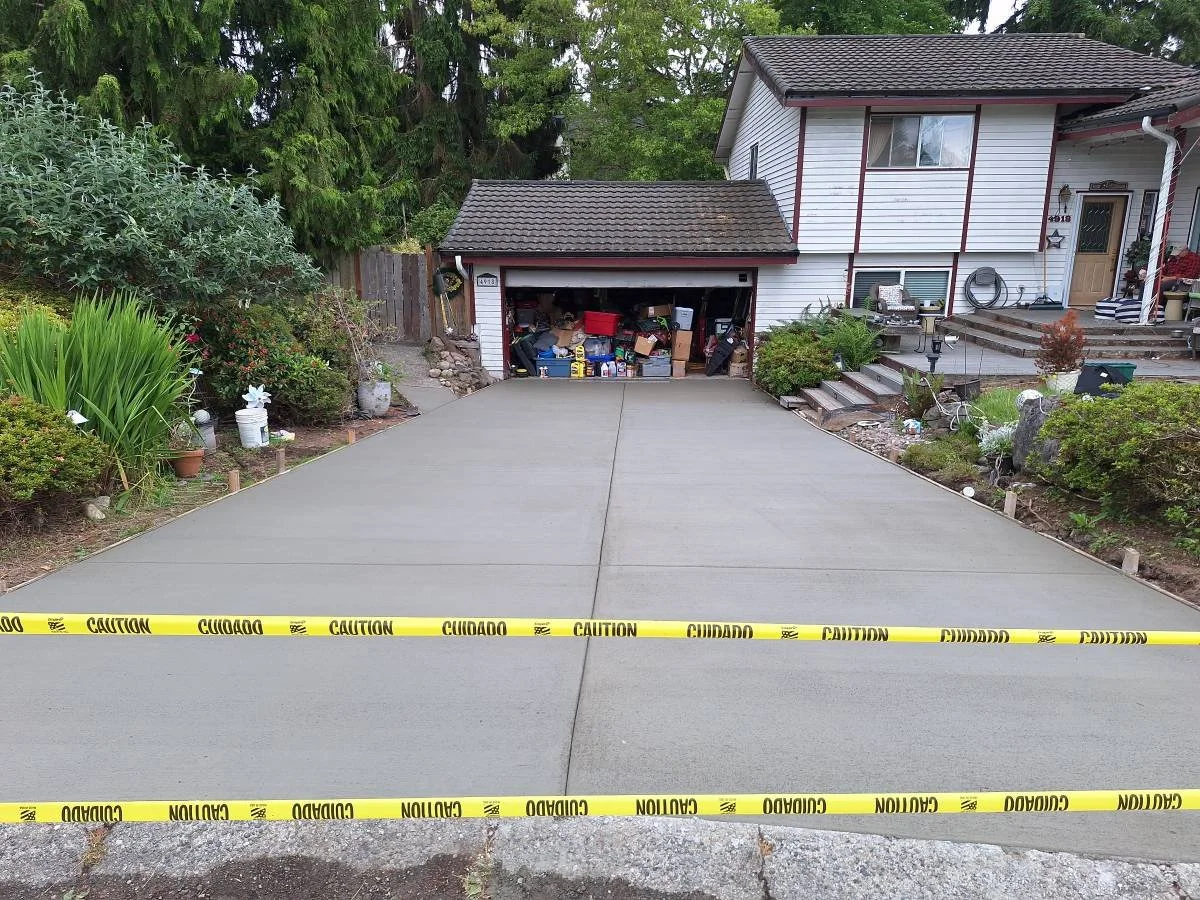 Newly poured concrete driveway in front of house, with yellow caution tape across it, and a garage filled with clutter and gardening supplies in the background.