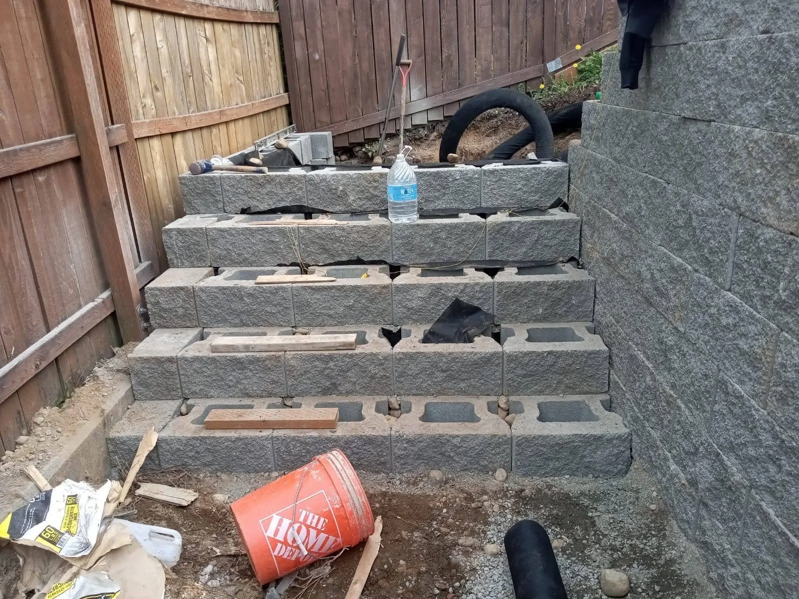 Construction site with stacked gray concrete blocks forming a small set of stairs, surrounded by tools, a water bottle, and construction debris, with a wooden fence and stone wall in the background.