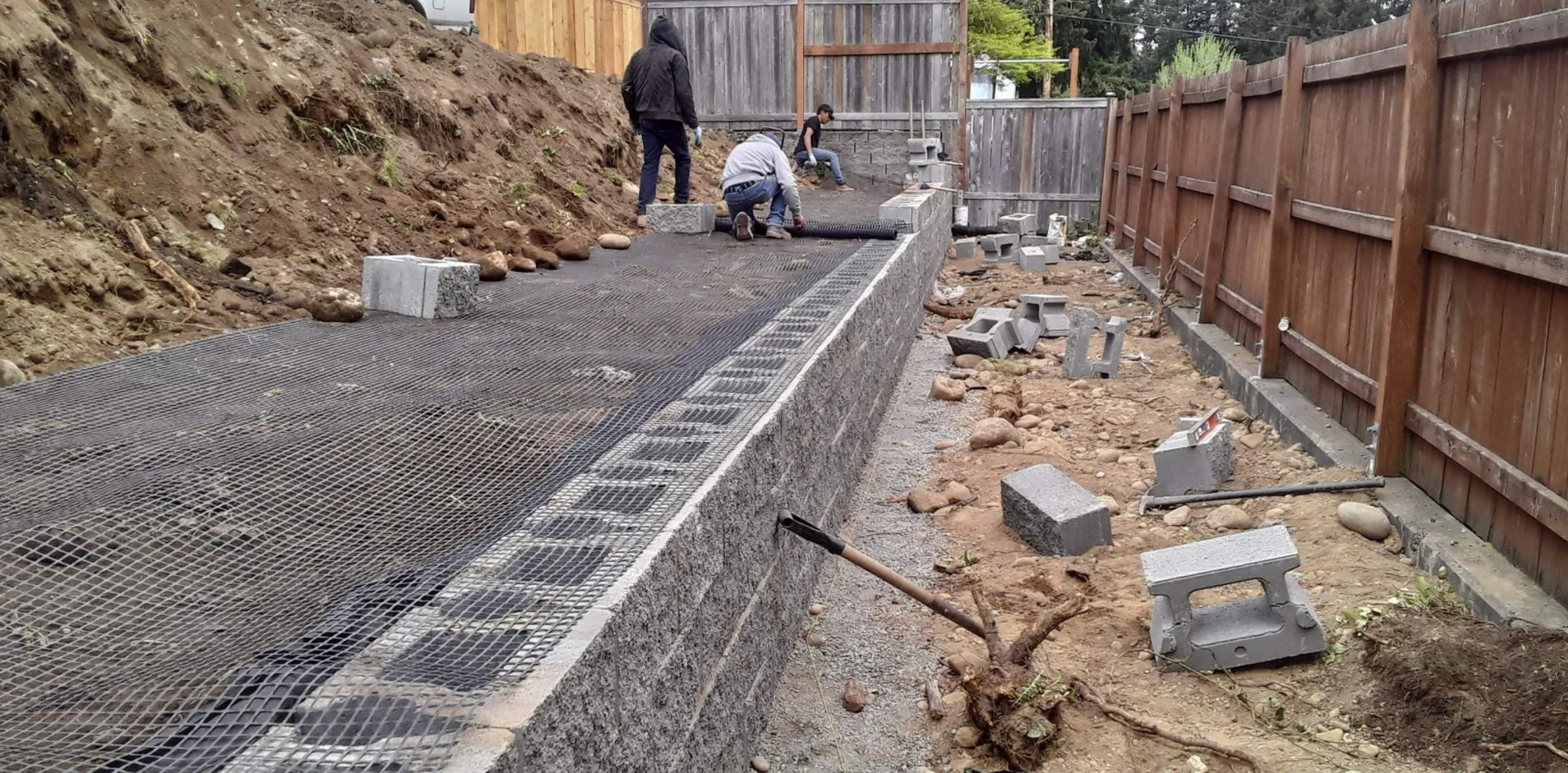 Construction workers installing a retaining wall with cinder blocks and metal mesh in a backyard, with a dirt hillside on one side and a wooden fence on the other.