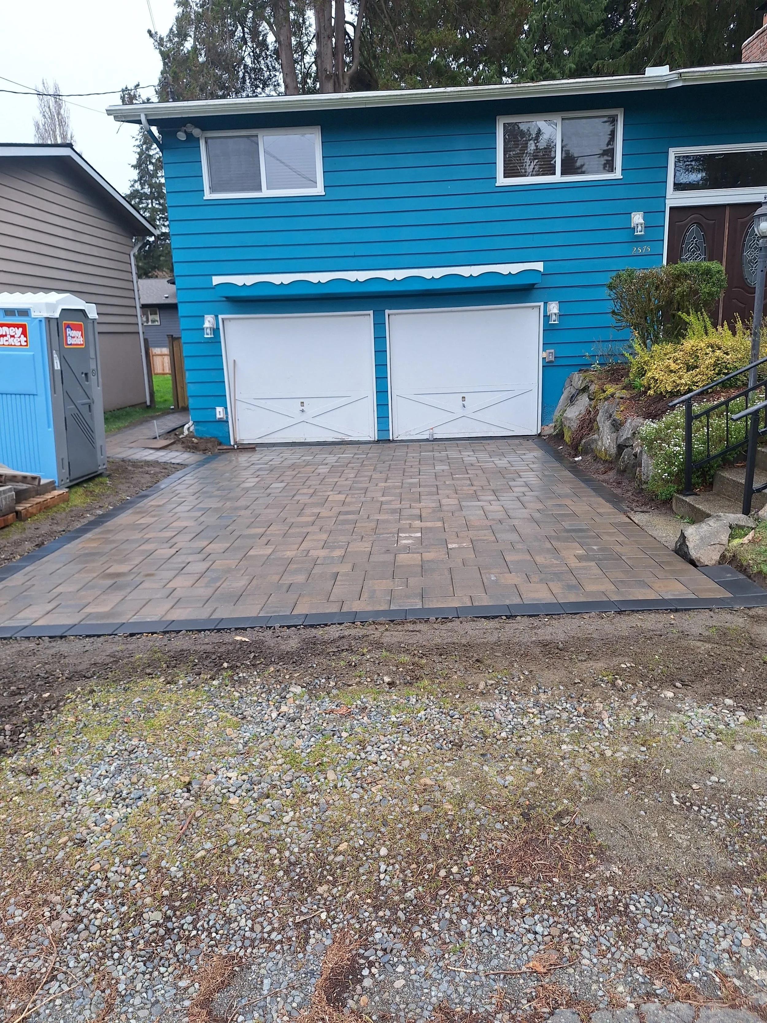 A two-car garage attached to a blue house with a brick pathway and landscaped plants on the right side. A Honey Bucket portable toilet is on the left side of the driveway, which is paved with brick pavers.