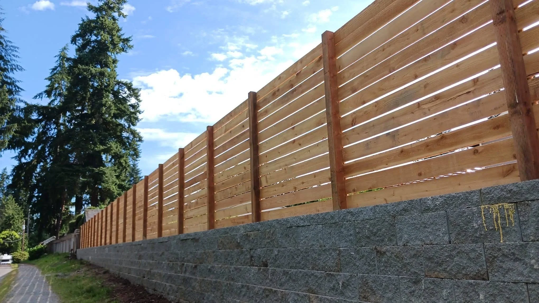 Wooden privacy fence installed on a stone retaining wall beside a sidewalk, with tall evergreen trees and a partly cloudy sky in the background.