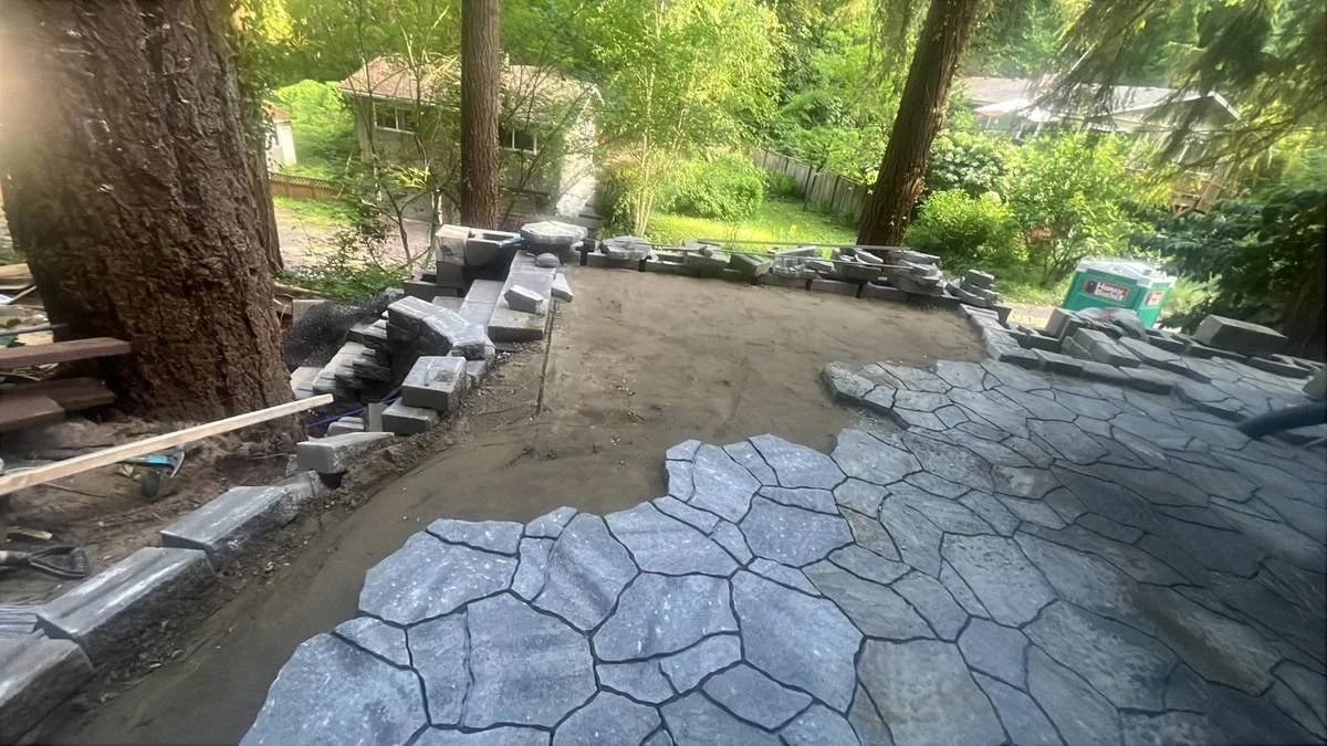 A backyard under construction with stone pathway installation, surrounded by tall trees and greenery, and a portable toilet in the background.