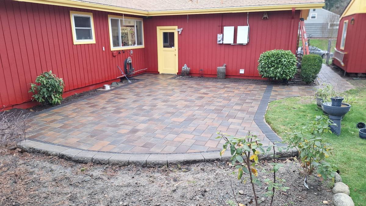 Backyard patio with brick pavers, red house with yellow door, garden bushes, and potted plants.