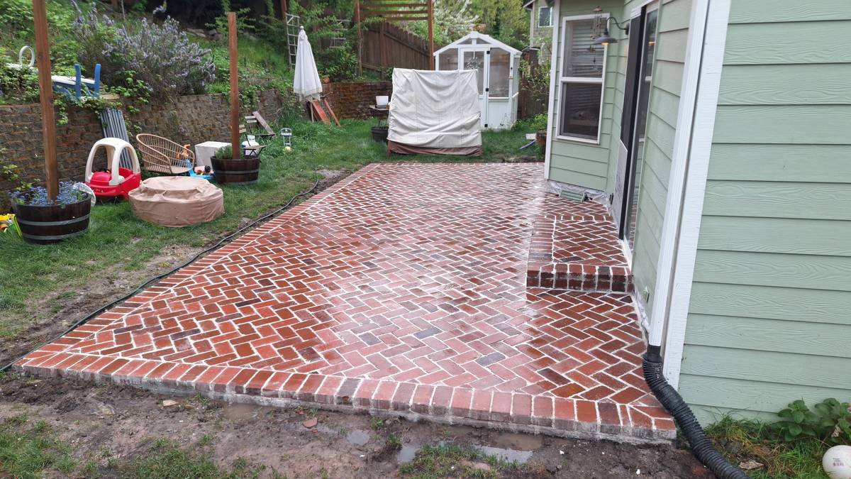 Newly constructed brick patio outside a house, with a small step leading up to a sliding door, surrounded by grass and garden items.