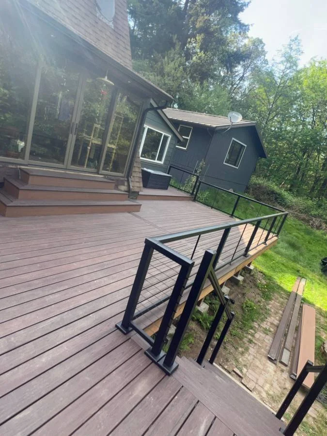 A wooden deck attached to a house with steps leading down to the yard, surrounded by forest, with black metal railing, a staircase, and a neighboring house visible in the background.
