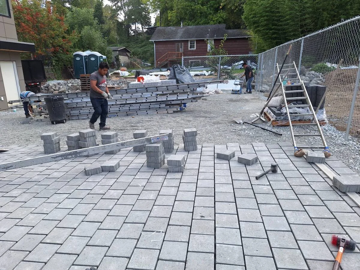 Construction workers installing grey paving stones on a driveway, with stacks of additional stones, tools, and a step ladder nearby.