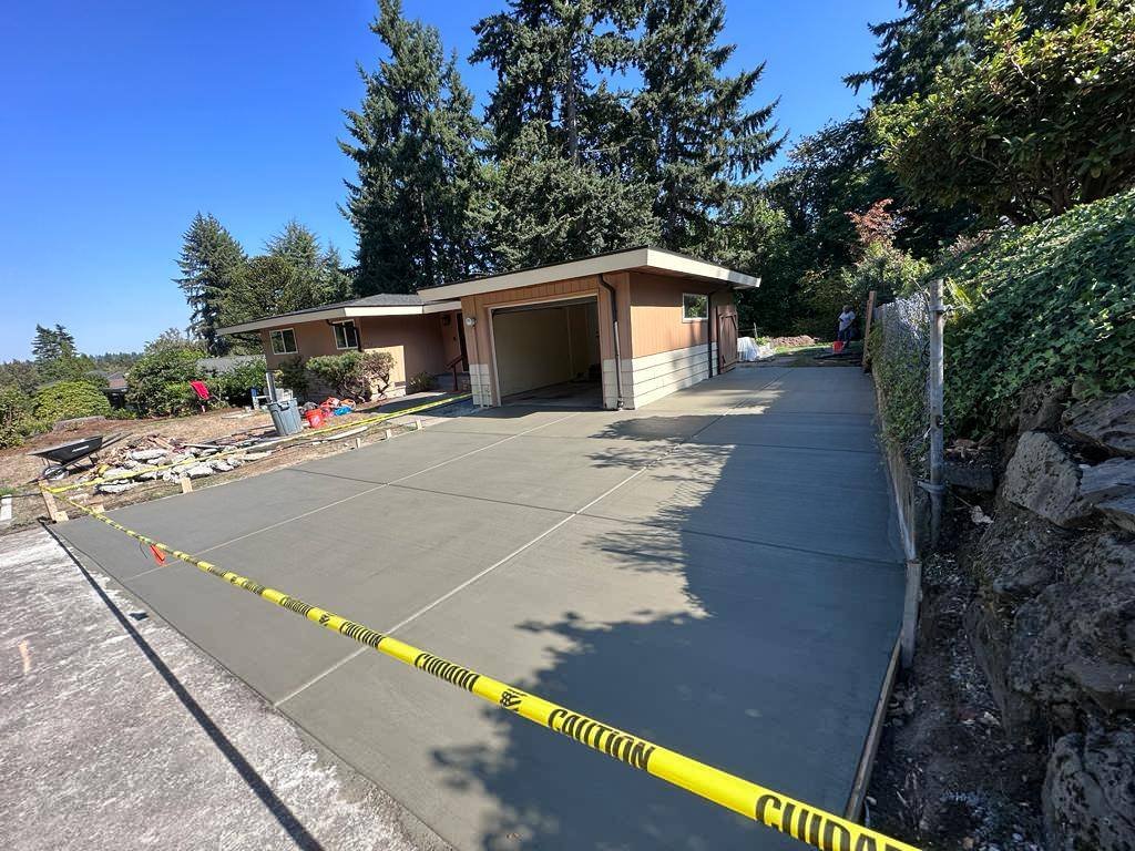 Newly poured concrete driveway and garage in front of a house, surrounded by caution tape, with trees and shrubs in the background.