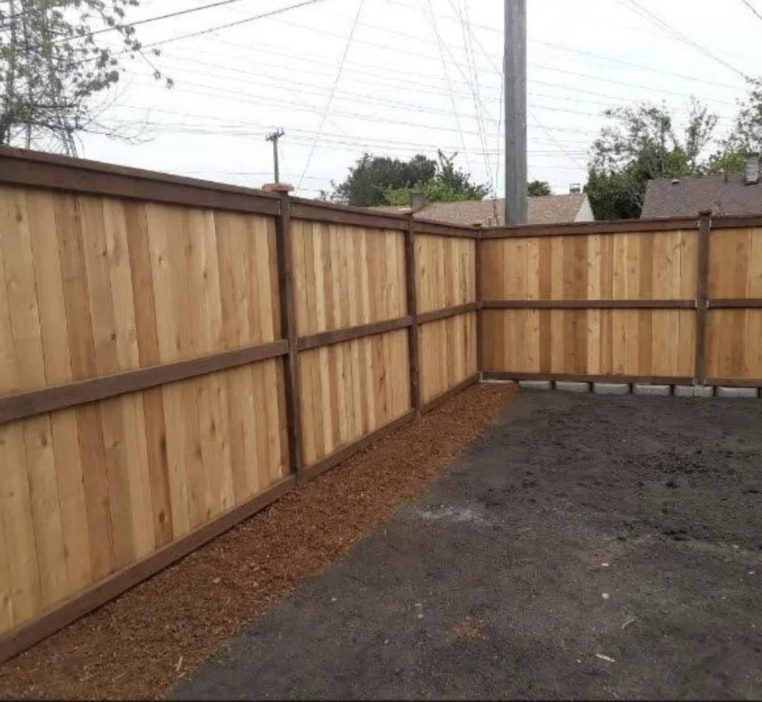 Newly installed wooden fence surrounding a backyard with dirt and gravel ground, and power lines in the background.