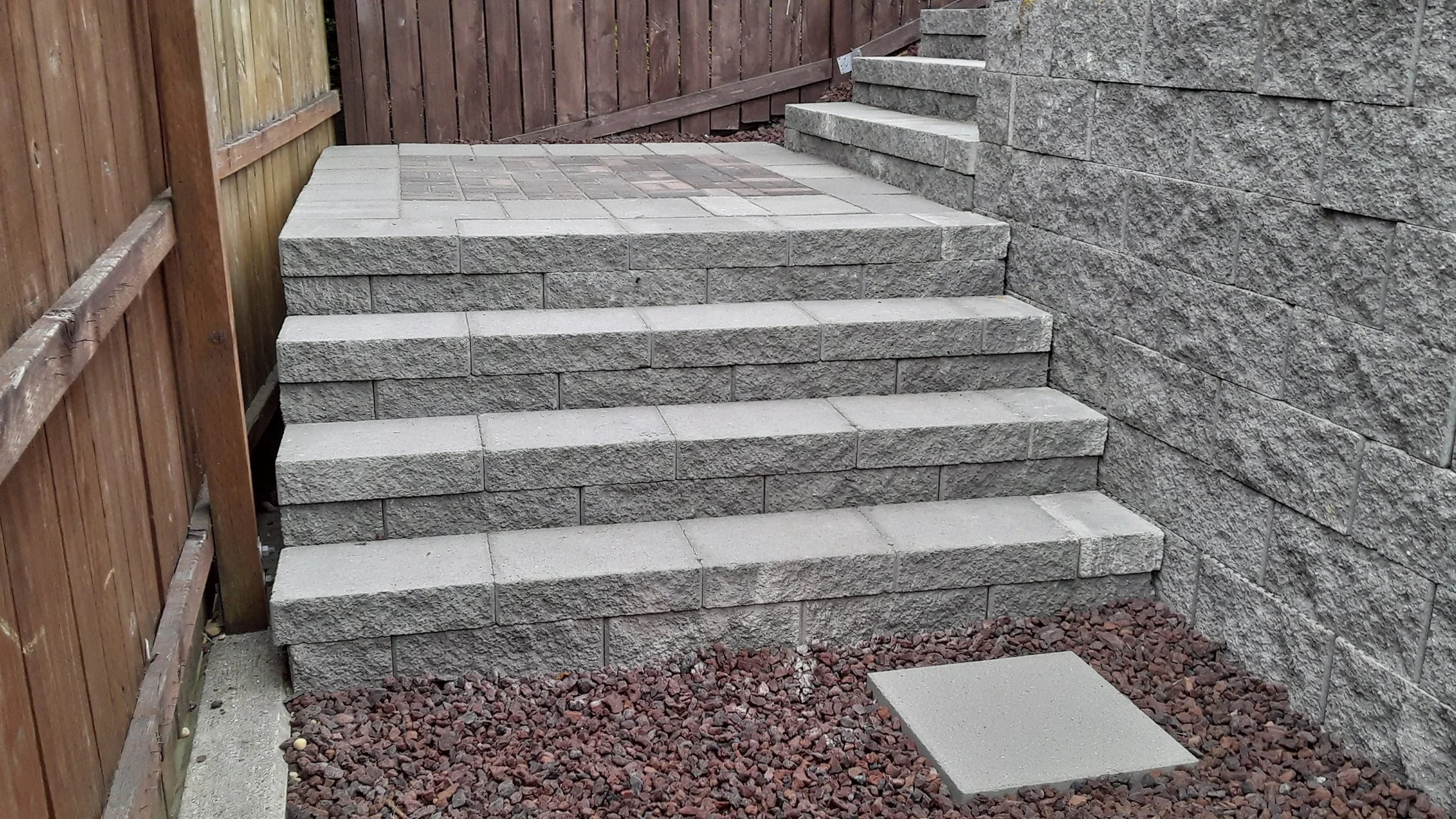 Concrete steps leading up to a higher level next to a stone wall, with a wooden fence on one side and red gravel around the base of the stairs.