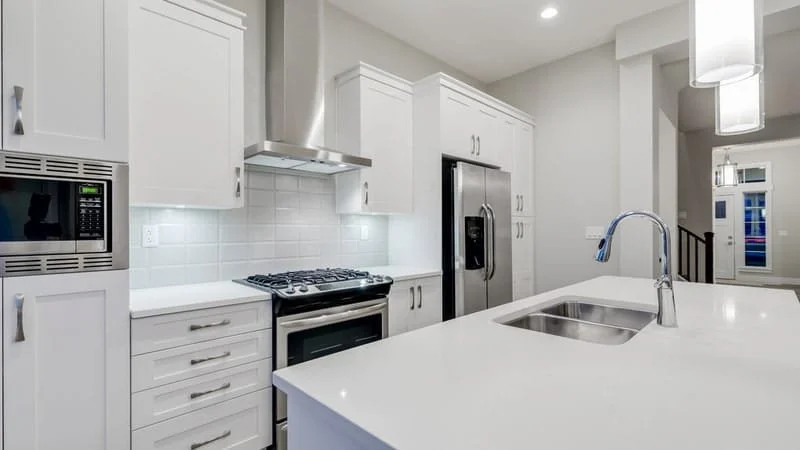 Modern white kitchen with appliances, including a microwave, refrigerator, stove, and oven, with a large sink and island counter.