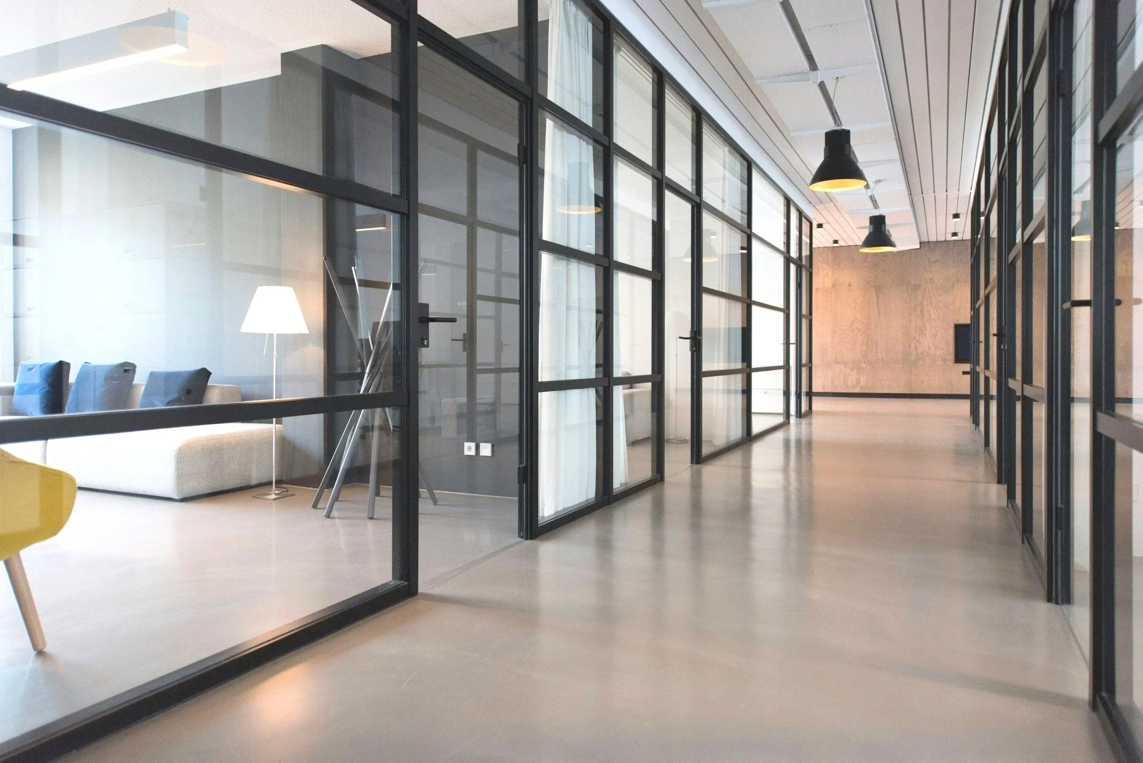 Modern office interior with glass walls, black metal frames, a sitting area with a white sofa, chairs, and a tall floor lamp, with a hallway and a large wall in the background.