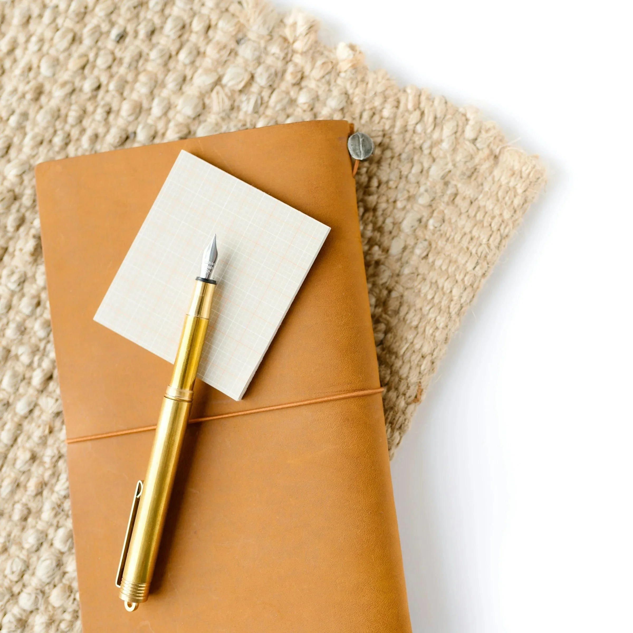 A composition of a brass pen, a small notepad, a brown leather folder, and a sheet of graph paper on a beige textured surface with a white background.