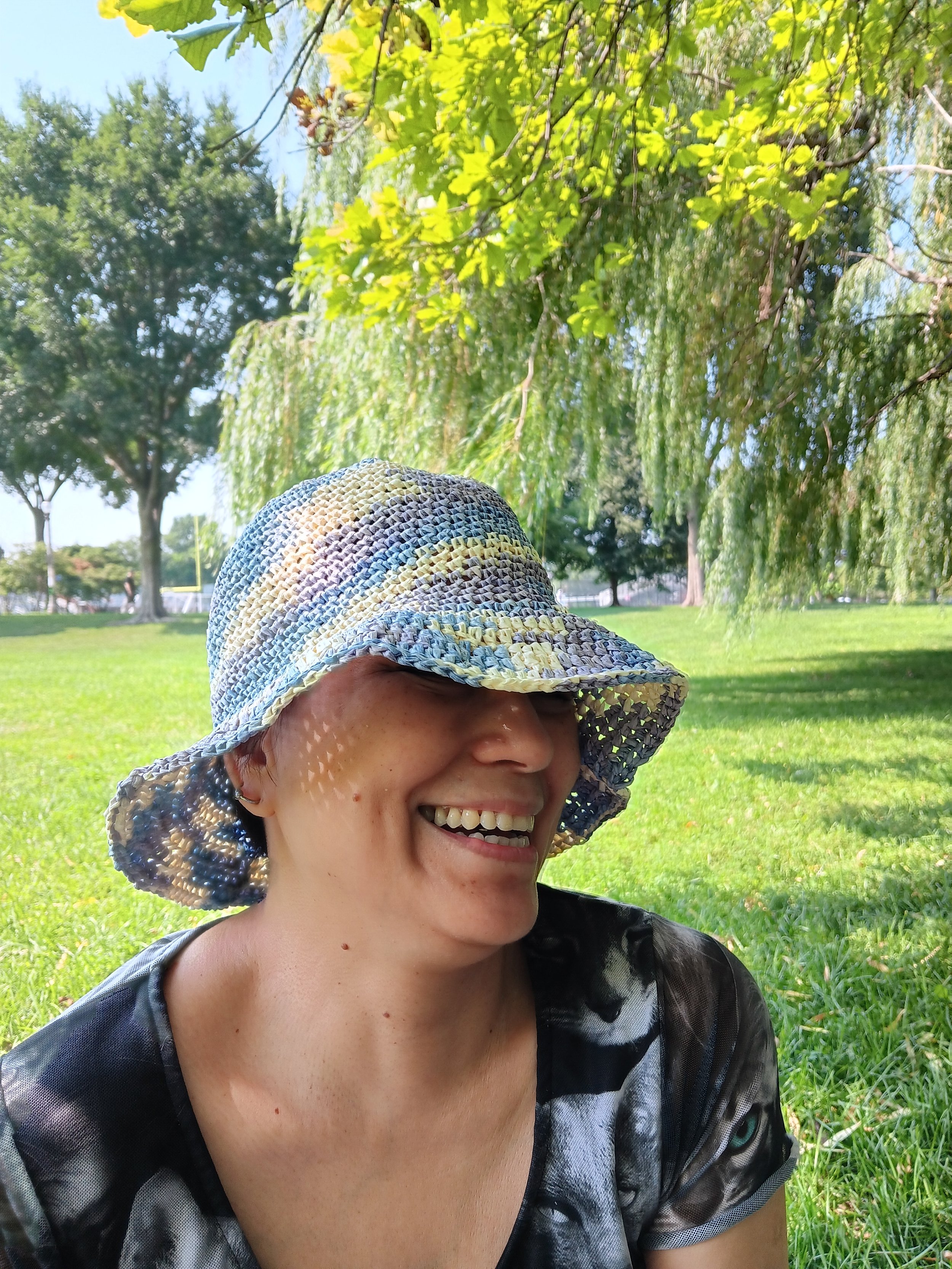 A woman smiling, wearing a multicolored woven sun hat, in an outdoor park with green grass and trees in the background.